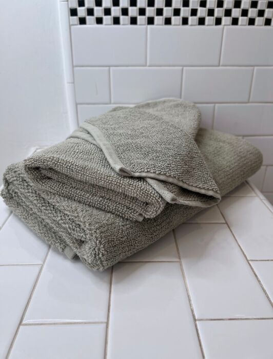 A neatly folded gray organic towel sits on a white tiled bathroom countertop with white subway tiles and a black-and-white mosaic accent in the background.