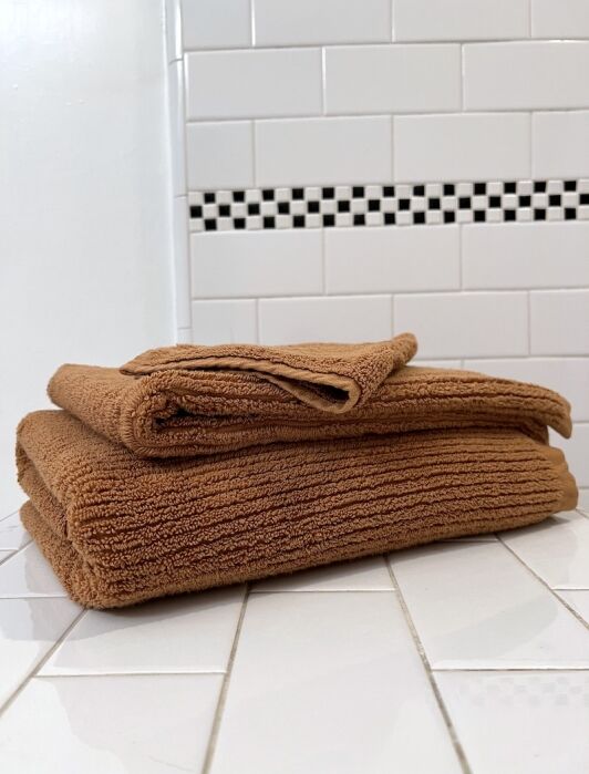 Two folded organic towels are stacked on a white tiled surface in front of a white tile wall with a black and white checkerboard accent.