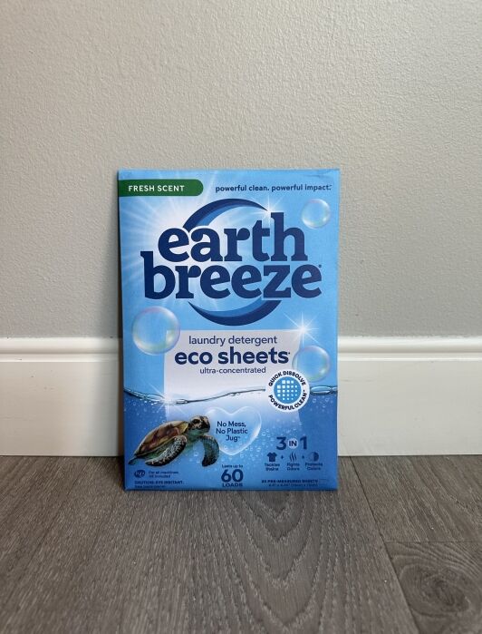 Box of Earth Breeze Fresh Scent eco sheets laundry detergent 3-in-1 placed upright against a grey wall on a wooden floor. The packaging features a turtle and various environmental benefits.