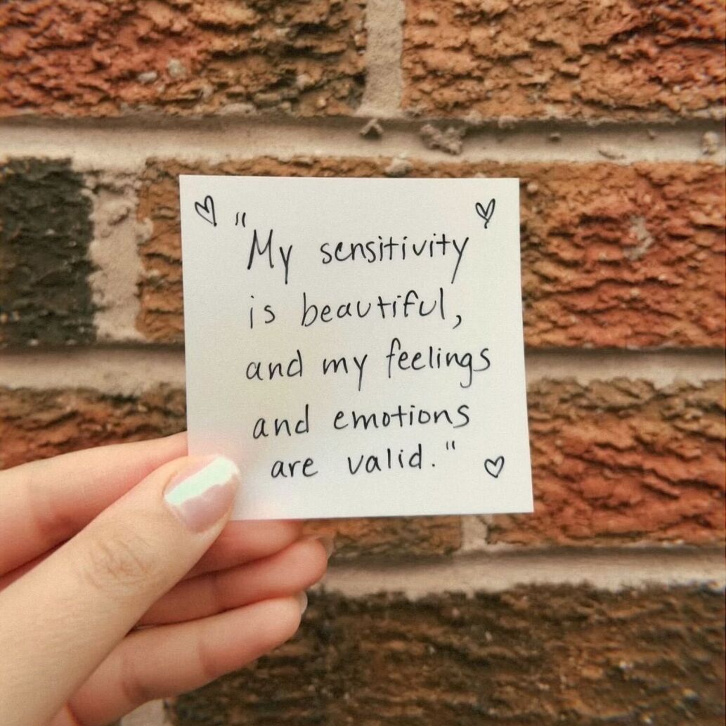 A sticky note held up in front of a brick wall, reading "My sensitivity is beautiful, and my feelings and emotions are valid".