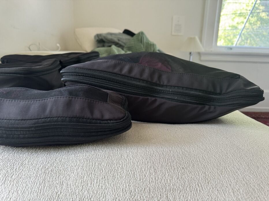 Two black zippered fabric bags are placed on a light-colored surface. A window and some clothing items are visible in the background.