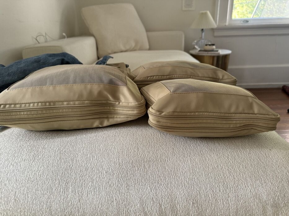 Three beige rectangular cushions are placed on a light-colored fabric surface, with a white chaise lounge and a small lamp in the background.