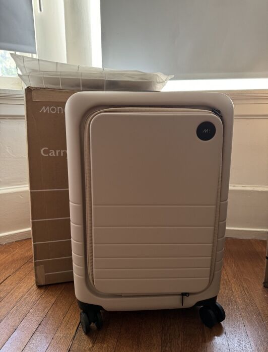 A light pink hard-shell carry-on suitcase stands on a wood floor in front of a cardboard box near a window.
