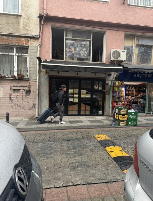Person walking down a street with a rolling suitcase in front of a small storefront, next to a shop with beverages on display outside.