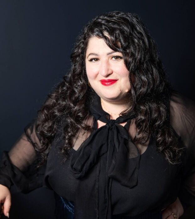 A person with long, curly dark hair wearing a sheer black blouse and red lipstick poses against a dark background.