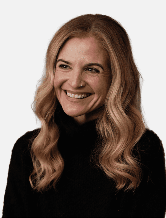 A woman with long wavy blonde hair, wearing a black turtleneck, smiles and looks slightly to the side against a plain background.