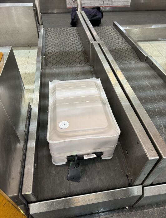 A white suitcase with a clear protective cover is placed on an airport baggage conveyor belt, with a tag attached to its handle.