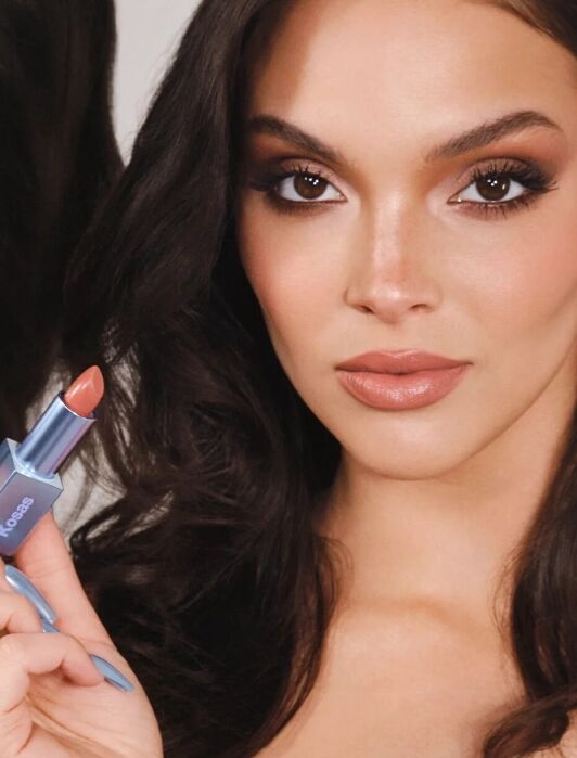 A person with long brown hair holds a lipstick tube. The text reads, "Kensington wears Weightless Lip Color in High Cut.