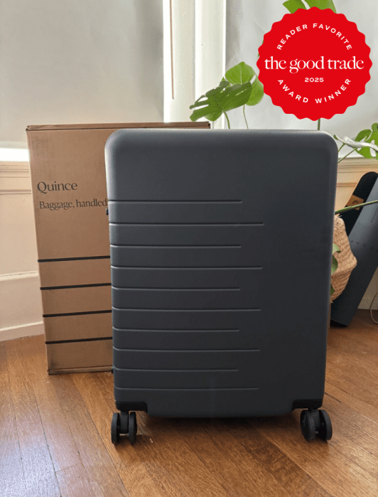 A dark gray suitcase with four wheels is in front of a brown box labeled 'Quince Baggage, handled.' A red award badge is in the corner.