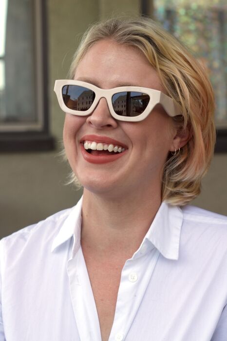 A person with short blonde hair wearing white-frame sunglasses and a white button-up shirt is smiling outdoors.