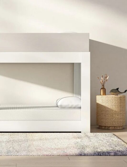 Minimalist white bunk bed with a ladder and shelves, placed in a bright room with light-colored walls, a gray rug, and a woven basket on the right. GREENGUARD Gold certification label on the upper left.