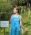 A person in a blue dress stands outdoors near a grassy area with trees. There's a sign nearby in French. The person has a tattoo on one arm and is looking to the side.