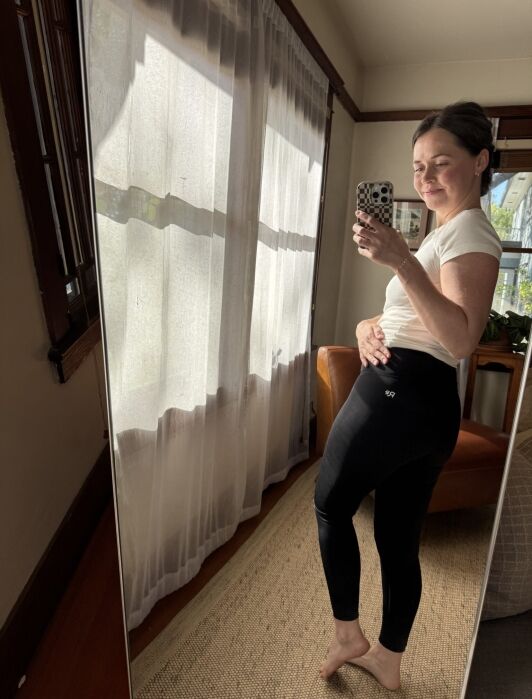 A person in a white t-shirt and black leggings stands barefoot in front of a mirror, taking a selfie in a sunlit room with curtains and a brown chair.