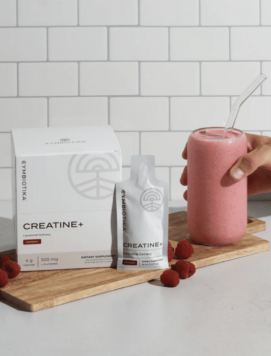 A hand holds a pink smoothie in a glass with a clear straw next to a creatine supplement box and packet on a wooden board, surrounded by raspberries against a tile background.
