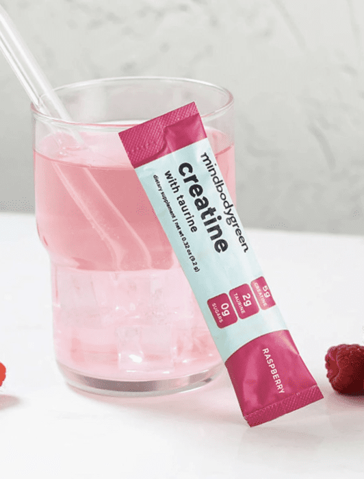 A glass of pink raspberry-flavored creatine drink with a clear straw, placed beside a creatine packet and fresh raspberries on a white surface.