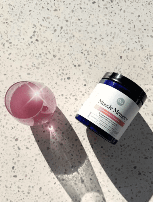 A glass of pink liquid beside a dark blue jar labeled "Muscle Memory." Both are placed on a speckled countertop with bright sunlight casting shadows.