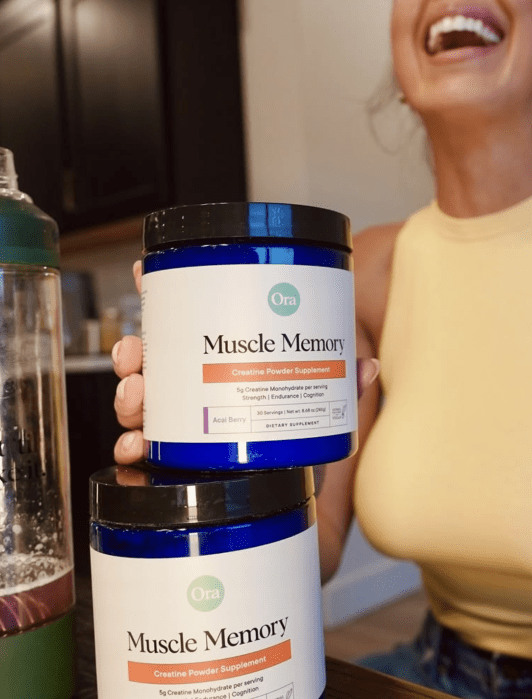 A woman in a yellow top holds a jar labeled "Muscle Memory." Another jar and a green drink bottle are on the table.