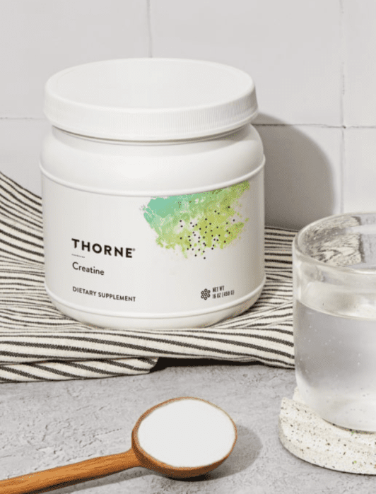 Container of Thorne creatine supplement with a wooden spoon of powder beside it and a glass of water on a coaster in the foreground.