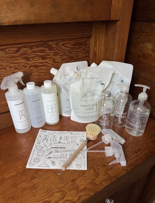 A collection of Common Good cleaning products, including bottles, refill pouches, glass containers, spray nozzles, a brush, and an illustrated instruction sheet on a wooden surface.