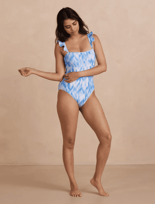 Person wearing a blue and white patterned one-piece swimsuit with ruffled straps stands against a plain beige background.