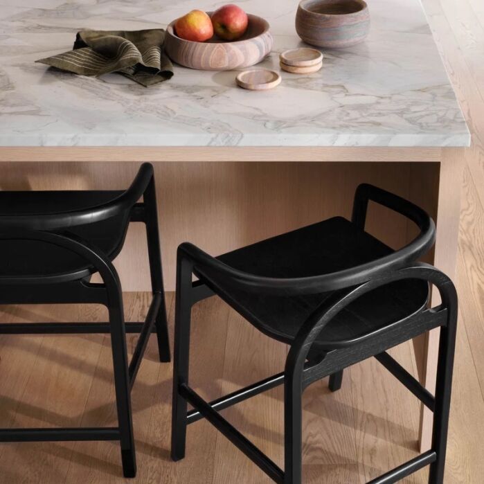 Two black stools are placed by a marble countertop with a wooden bowl of apples, a cloth napkin, and two small lidded containers on top.