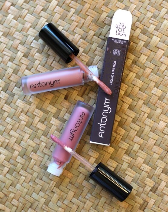 Two open tubes of Antonym liquid lipstick with applicators and a branded box are arranged on a woven surface.