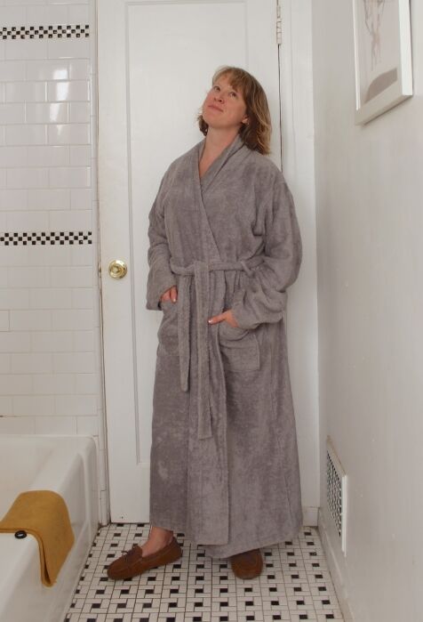 A person wearing a gray bathrobe and brown slippers stands in a white-tiled bathroom, looking up with a neutral expression.