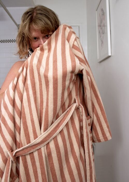 A person stands in a bathroom, partially covering their face with a beige and white striped bathrobe.