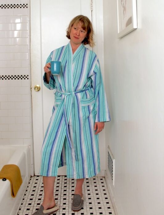 Person standing in a bathroom wearing a blue and white striped robe and slippers, holding a blue mug, with a towel on the bathtub and framed art on the wall.