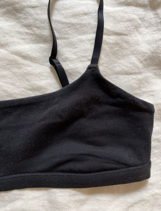 A black one-shoulder bralette with a single strap is laid flat on a light-colored fabric background.