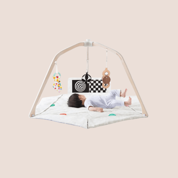 A baby lies on a play mat under an activity gym with hanging toys, including a mirror, wooden rings, and high-contrast cards.