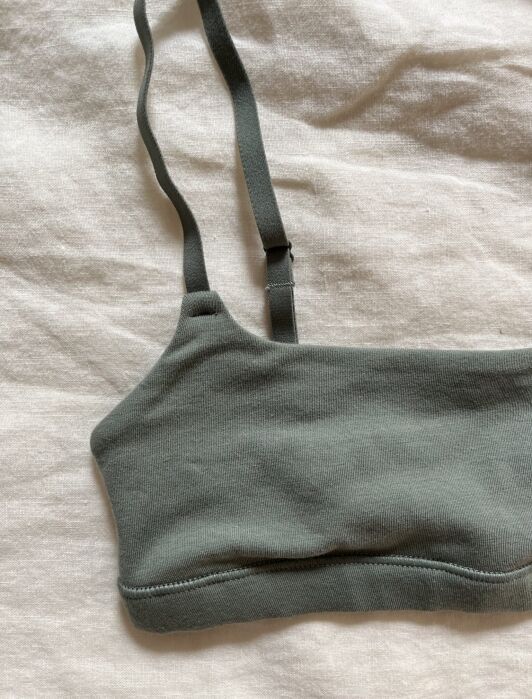A close-up of a green, ribbed bralette with adjustable straps lying on a textured white fabric background.