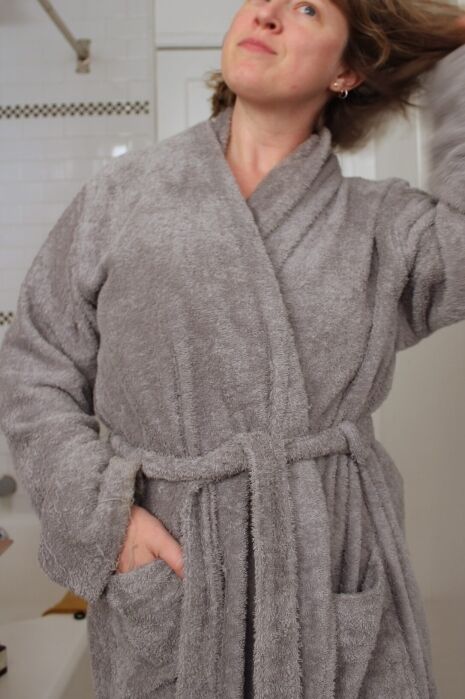 A person wearing a gray bathrobe stands in a bathroom, looking up and touching their hair.