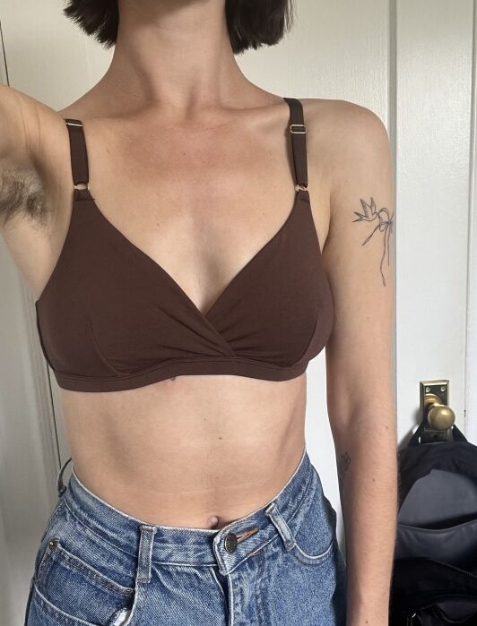 A person wearing a brown bralette and blue jeans stands indoors. Their left arm is raised, showing an armpit and a floral tattoo on their upper arm.