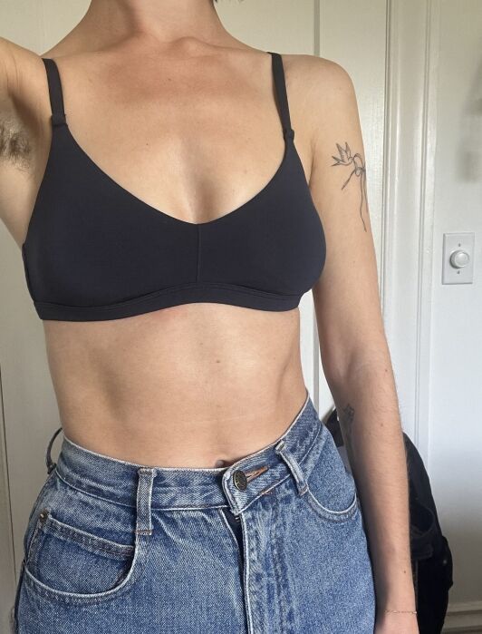 A person wearing a black bralette and high-waisted blue jeans stands indoors. Their left arm shows a minimalist flower tattoo.