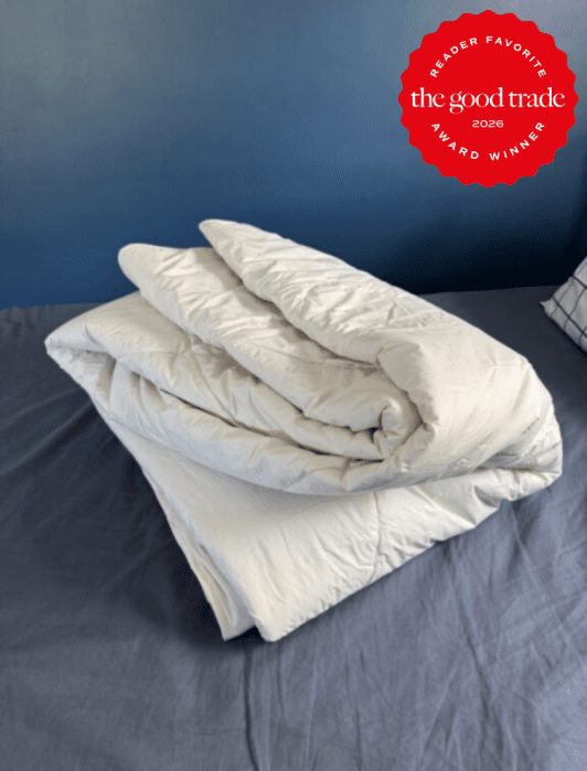A folded white comforter sits on a bed with a blue wall and a plaid pillow in the background. A red badge in the corner reads, “The Good Trade Award Winner 2026.”.