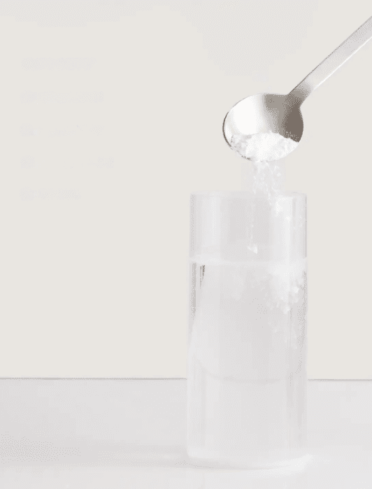 A spoon pours a white powder, likely sugar or salt, into a clear glass of water against a plain light background.