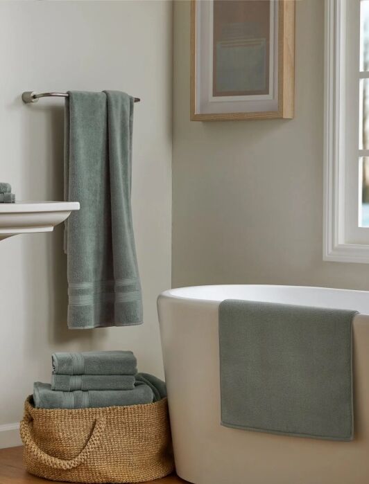 A bathroom with a white tub, a sage green organic towel draped over it, a matching towel on a rack, folded organic towels in a woven basket, and a framed picture on the wall.