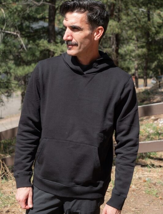Man with a mustache wearing a black hoodie stands outdoors in a wooded area, looking to the side.