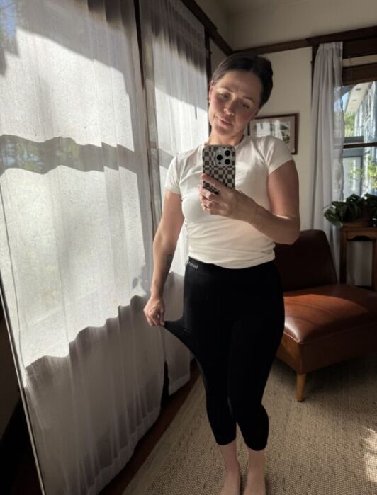 A person stands in a sunlit room, wearing a white shirt and black leggings, holding a phone for a mirror selfie and pulling the waistband of their leggings to show looseness.