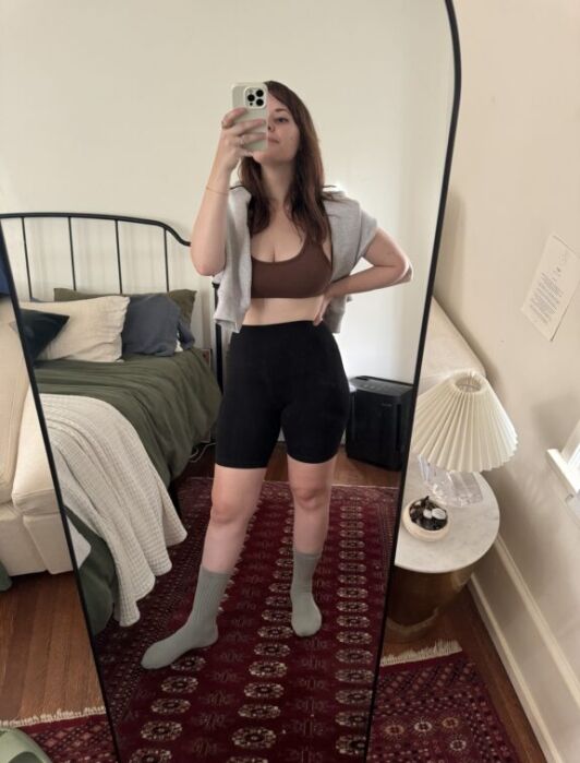 A woman stands in front of a full-length mirror taking a selfie, wearing a brown sports bra, black shorts, gray socks, and a gray top draped over her shoulders in a bedroom.