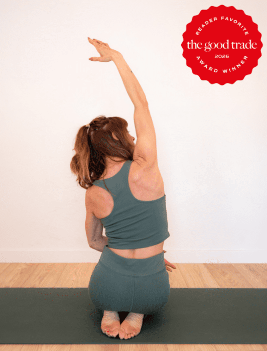 A woman in green workout attire sits on a yoga mat performing a seated side stretch; a red "The Good Trade Award Winner 2026" badge is in the top right corner.