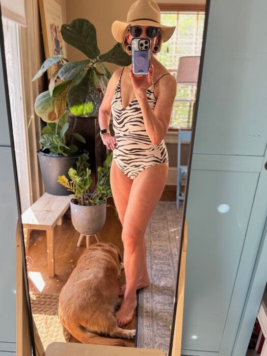 Person wearing a zebra-patterned swimsuit and hat takes a mirror selfie in a room with plants; a brown dog lies on the rug by their feet.