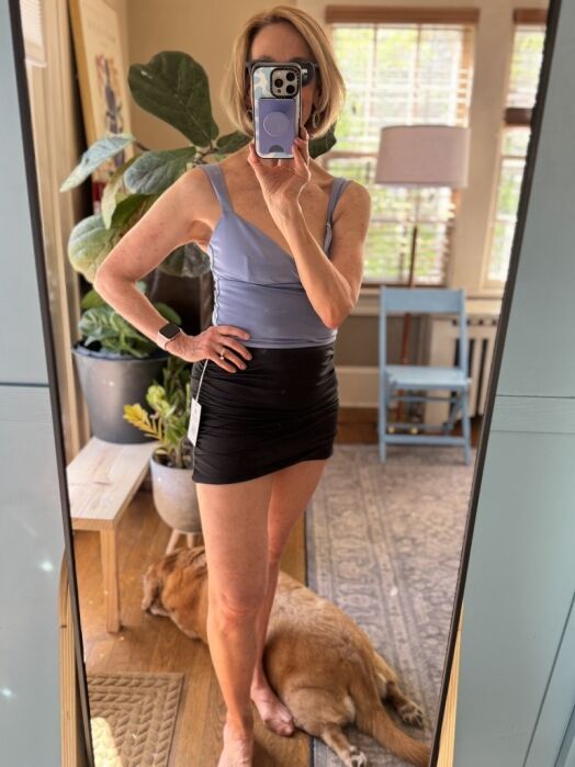 A woman takes a mirror selfie wearing a lavender top and black skirt. A dog lies on the floor behind her in a well-lit room with plants and furniture.
