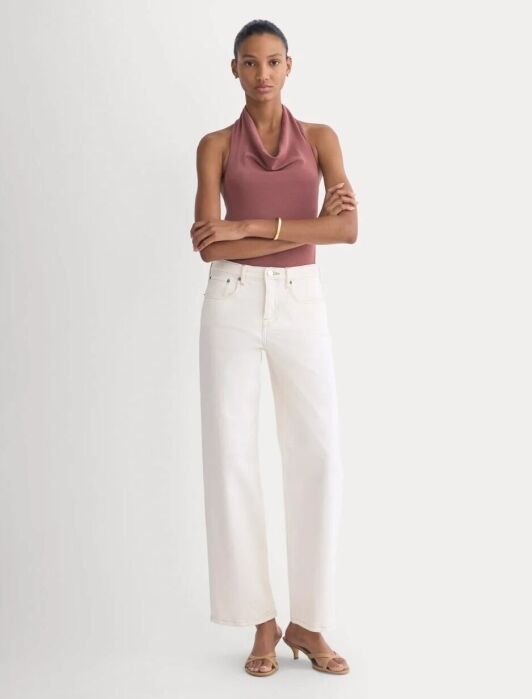A woman stands with arms crossed, wearing a mauve sleeveless cowl-neck top, white wide-leg jeans, beige heeled sandals, and a gold bracelet against a plain background.