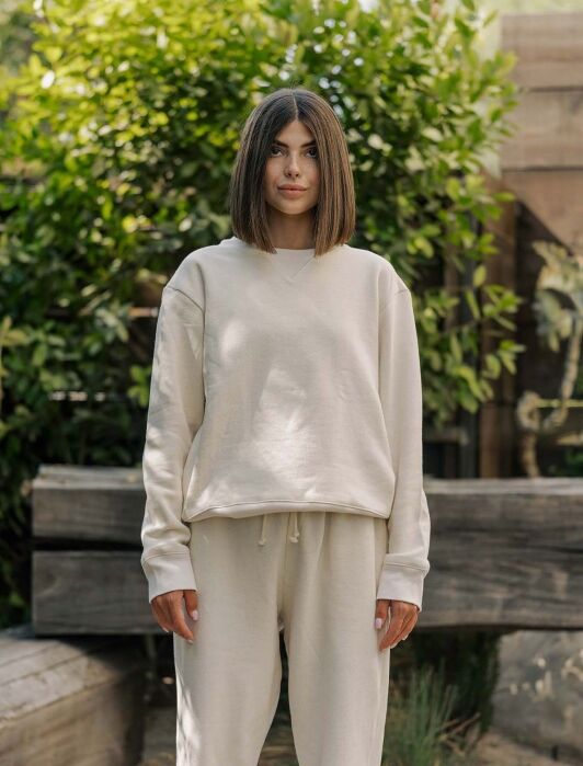 A person with straight, shoulder-length hair stands outdoors wearing a loose-fitting, light beige sweatshirt and matching sweatpants. Green foliage and wooden beams are in the background.