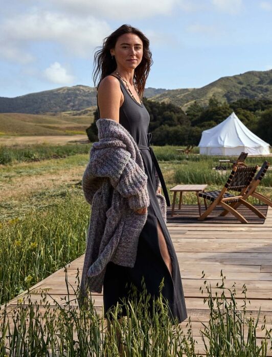 Woman in a long black dress and gray cardigan stands on a wooden deck in a grassy outdoor area with tents and mountains in the background.