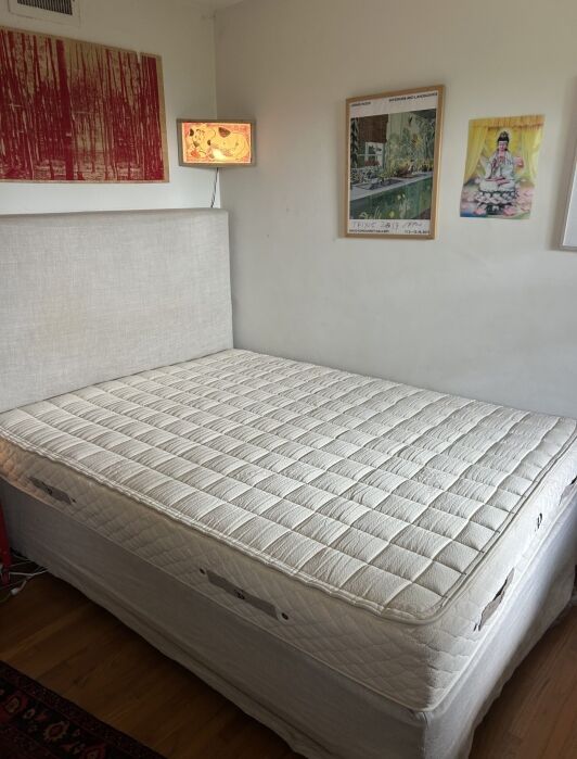 A neatly made beige bed with no sheets or pillows, placed against a white wall with three colorful artworks hanging above it.