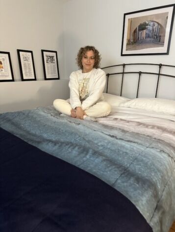A person with curly hair sits cross-legged on a bed in a neatly decorated bedroom with framed art on the wall.