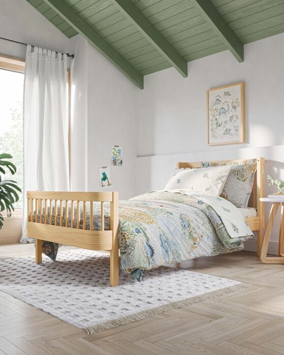 A neatly made wooden bed with patterned bedding sits on a white rug in a bright bedroom with green ceiling beams, a window with sheer curtains, and framed art on the wall.